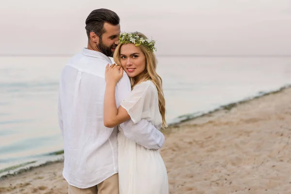 Sposo abbracciare sposa attraente in ghirlanda e lei guardando la fotocamera sulla spiaggia — Foto stock