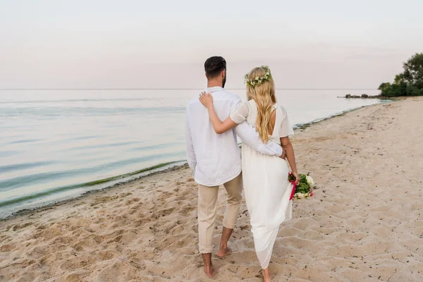 Visão traseira da noiva com buquê de casamento e noivo abraçando e andando na praia — Fotografia de Stock