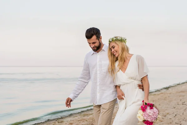 Handsome groom hugging beautiful bride with wedding bouquet and they walking on beach — Stock Photo