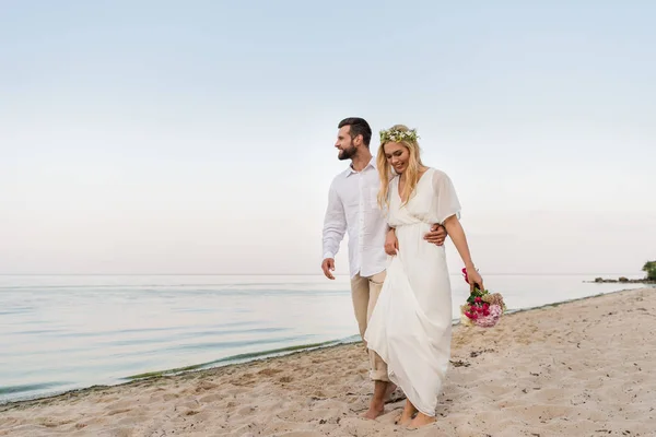 Schöner Bräutigam umarmt schöne Braut mit Brautstrauß und sie zu Fuß am Meer — Stockfoto