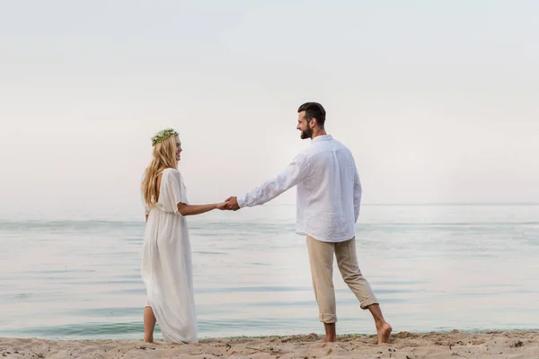 Vista trasera de la novia y el novio tomados de la mano y mirándose unos a otros cerca del océano - foto de stock