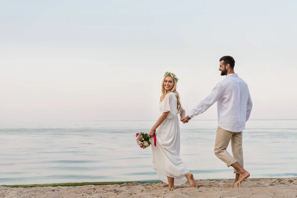 Happy bride and groom holding hands and walking with wedding bouquet on beach — Stock Photo
