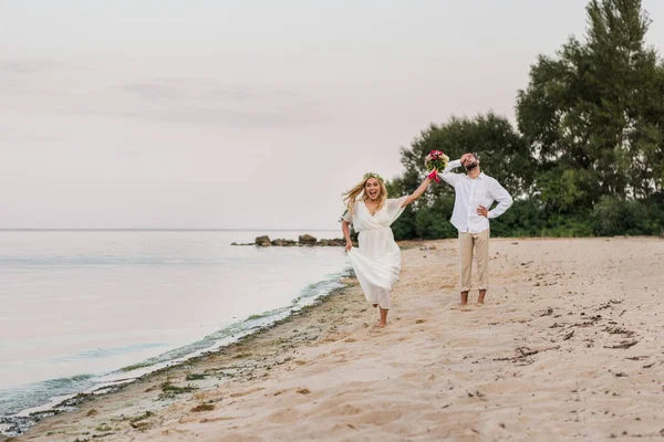 Щаслива наречена біжить з весільним букетом, наречений посміхається на пляжі — стокове фото