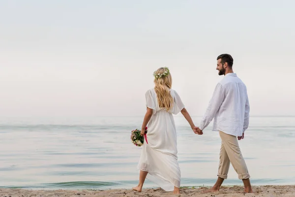 Visão traseira da noiva e do noivo de mãos dadas e andando com buquê de casamento na praia — Fotografia de Stock