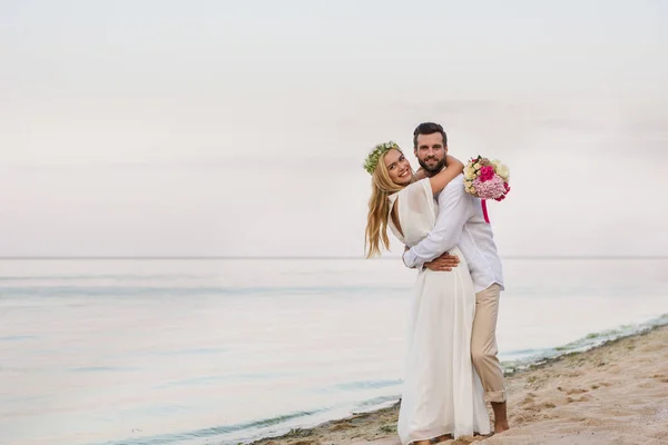 Noiva feliz e noivo abraçando na praia e segurando buquê de casamento — Fotografia de Stock