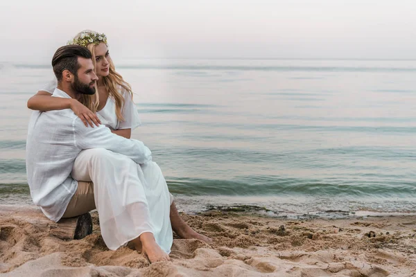 Elegante noivo e noiva sentado no log na praia e olhando para longe — Fotografia de Stock