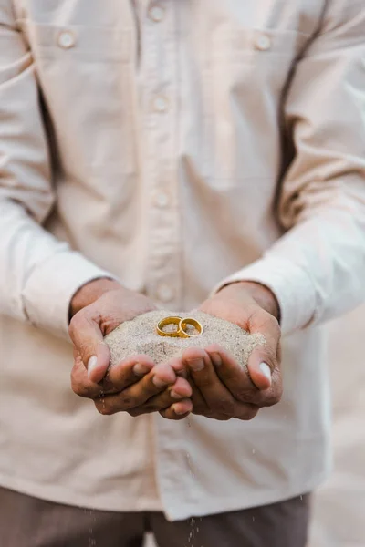 Cropped image of groom holding wedding rings with sand in hands on beach — Stock Photo