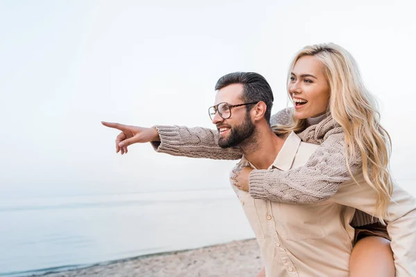 Boyfriend giving piggyback to smiling girlfriend in autumn outfit and she pointing on something on beach — Stock Photo