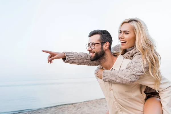 Novio dando piggyback a la novia sonriente en traje de otoño y ella señalando algo en la playa - foto de stock