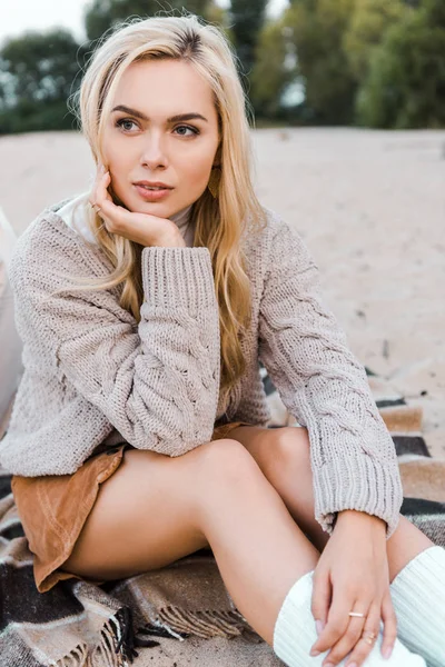 Jolie fille blonde coûteuse en tenue d'automne assis sur la plage et regardant loin — Photo de stock