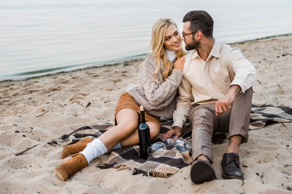 Casal elegante em roupa de outono olhando uns para os outros na praia com garrafa de vinho tinto — Fotografia de Stock