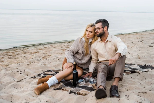 Stylish couple in autumn outfit sitting on beach with bottle of red wine — Stock Photo