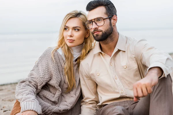 Pareja elegante en traje de otoño sentado en la playa y mirando hacia otro lado - foto de stock