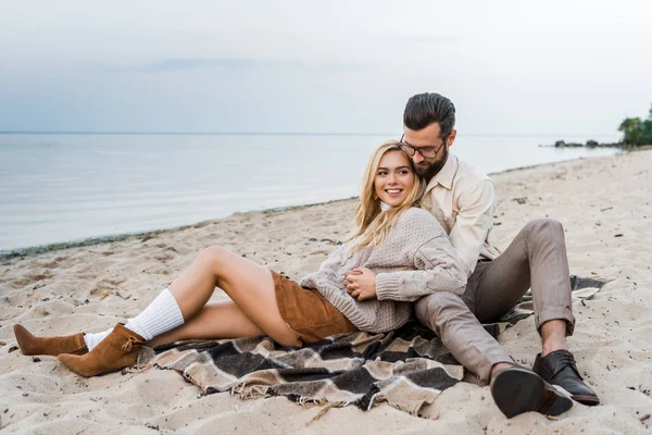 Sonriente pareja en otoño traje sentado en manta y abrazos en la playa - foto de stock