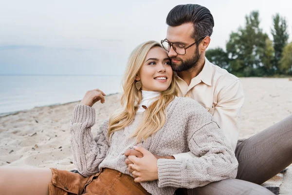 Smiling girlfriend and boyfriend in autumn outfit sitting and hugging on beach — Stock Photo