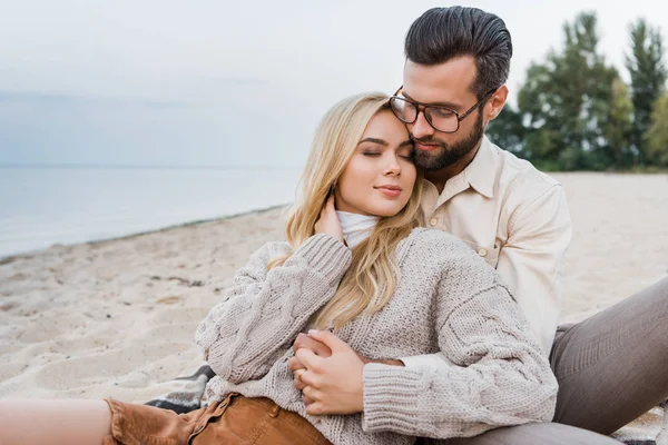 Pareja cariñosa en traje de otoño sentado y abrazándose en la playa - foto de stock