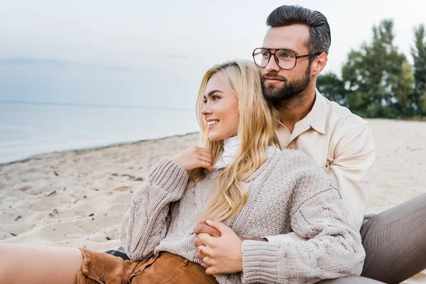 Fröhliches Paar im Herbst-Outfit sitzt und umarmt sich am Strand — Stockfoto