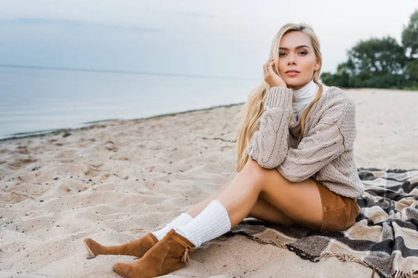 Attraktive blonde Mädchen im Herbst-Outfit am Strand sitzend, das Kinn auf der Hand liegend und in die Kamera blickend — Stockfoto