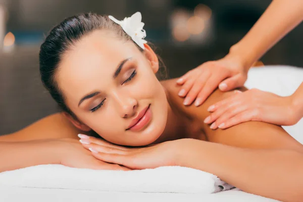 Hermosa joven con flor en el pelo teniendo masaje en el salón de spa - foto de stock