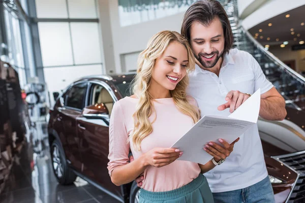 Portrait of cheerful couple with catalog buying car at dealership salon — Stock Photo