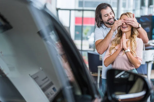 Homem sorridente surpreendente namorada com carro novo no salão de concessionárias — Fotografia de Stock