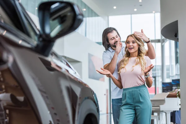 Smiling man surprising girlfriend with new car at dealership salon — Stock Photo