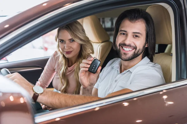 Sorrindo casal com chave de carro sentado em carro novo no salão de concessionárias — Fotografia de Stock