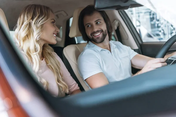 Sonriente pareja mirando el uno al otro en el nuevo automóvil en el salón de concesionarios - foto de stock