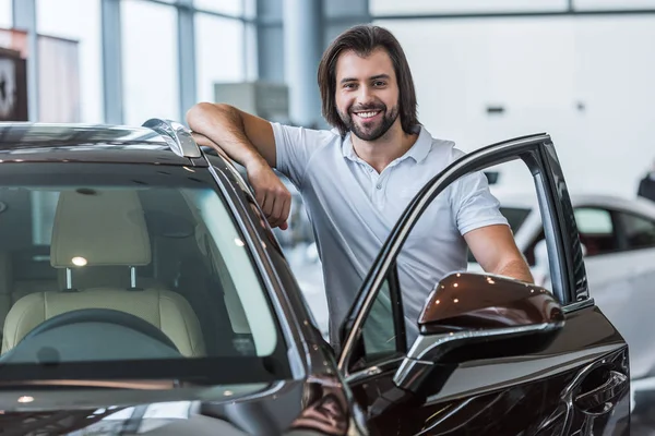 Портрет улыбающегося мужчины, стоящего у новой машины в салоне автосалона — стоковое фото