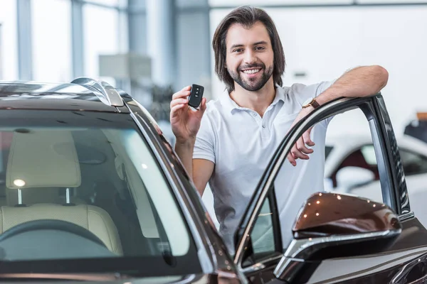 Ritratto dell'uomo felice con chiave dell'automobile che si leva in piedi al nuovo automobile nel salone della concessionaria — Foto stock