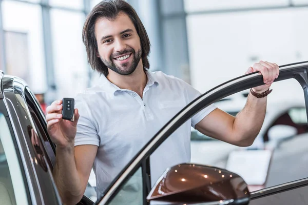 Retrato de homem feliz com chave de carro em pé no carro novo no salão de concessionárias — Fotografia de Stock