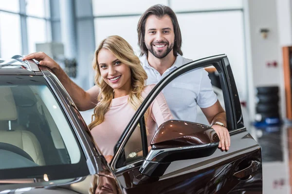 Retrato de la pareja sonriente de pie en el coche nuevo en el salón de concesionarios - foto de stock