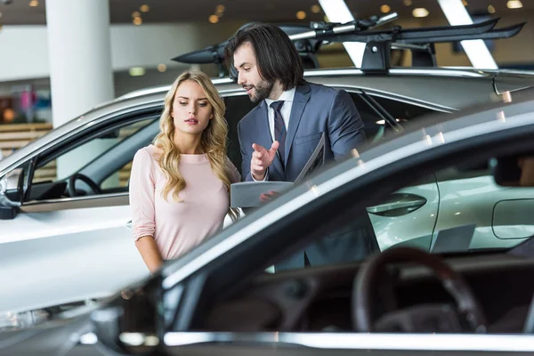 Verkäufer in festlicher Kleidung empfiehlt Frau im Autohaus das Auto — Stockfoto