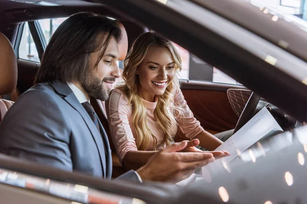 Vendedor de salão de concessionária e cliente sorridente feminino sentado no carro no salão de auto — Fotografia de Stock