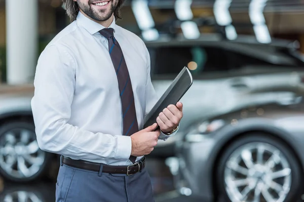 Cropped shot of seller with folder standing in dealership salon with cars on background — Stock Photo