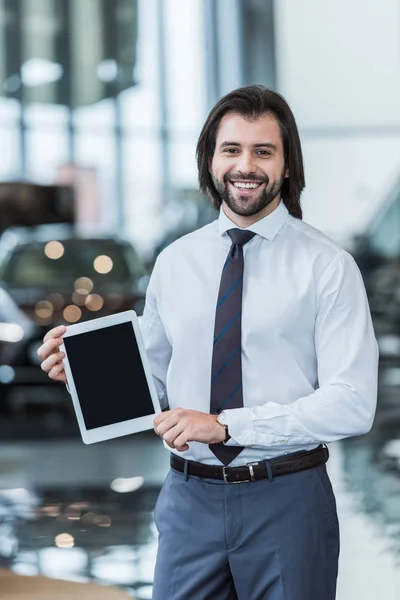 Ritratto del venditore maschio sorridente in abito formale che mostra tablet con schermo bianco al salone di concessionaria — Foto stock