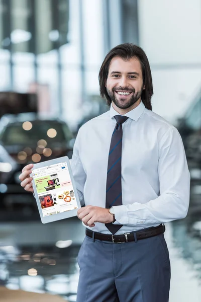 Smiling dealership salon seller in formal wear showing tablet with ebay website on screen in hands — Stock Photo