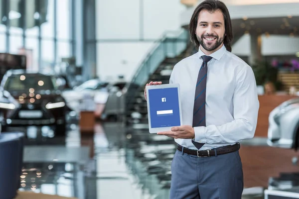 Portrait of cheerful dealership salon seller in formal wear showing tablet with facebook logo on screen in hands — Stock Photo