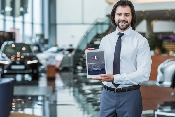 Portrait of cheerful dealership salon seller in formal wear showing tablet with tumblr logo on screen in hands — Stock Photo