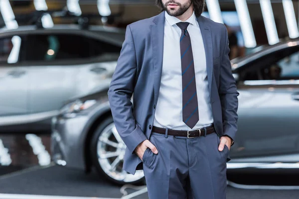 Vue partielle de l'homme d'affaires en costume élégant debout dans le salon de concession avec de nouvelles voitures sur fond — Photo de stock