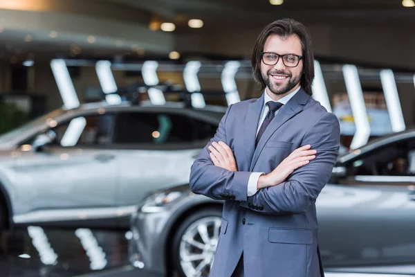 Улыбающийся бизнесмен с скрещенными руками, стоящий в салоне дилеров с новыми автомобилями на заднем плане — стоковое фото