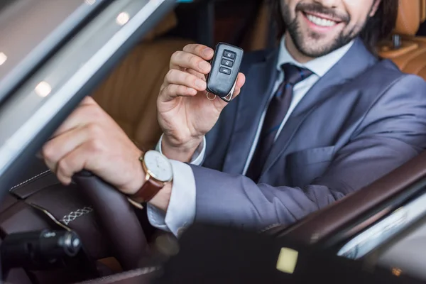 Vista parziale di un uomo d'affari sorridente con chiave della macchina in mano seduto in un'auto nuova nel salone di concessionaria — Foto stock