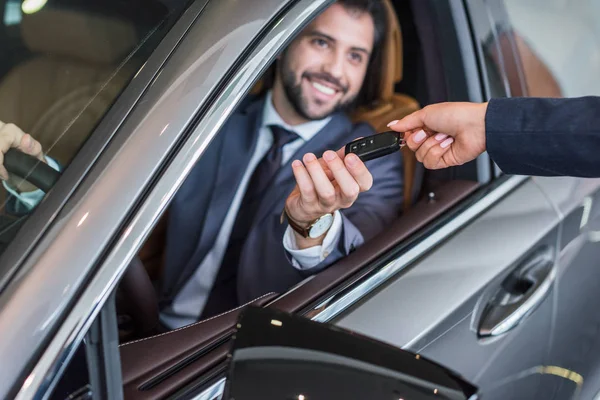 Partial view of female seller giving car key to smiling businessman in new car at dealership salon — Stock Photo