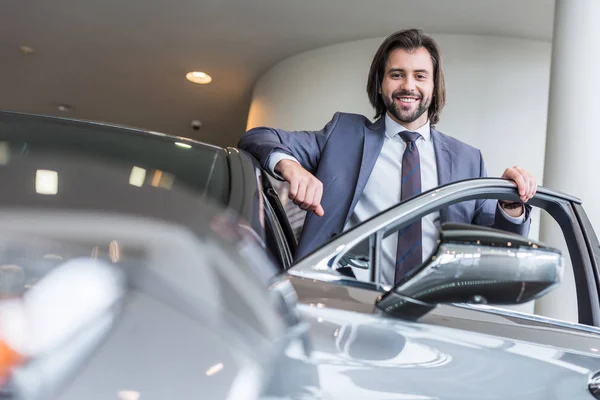 Happy stylish businessman standing at new car at dealership salon — Stock Photo