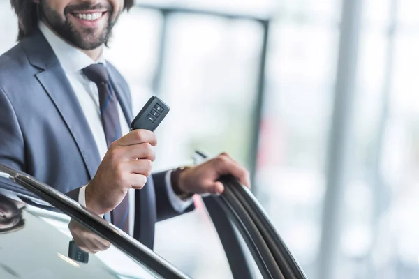 Cropped shot of smiling businessman with car key standing at new car at dealership salon — Stock Photo