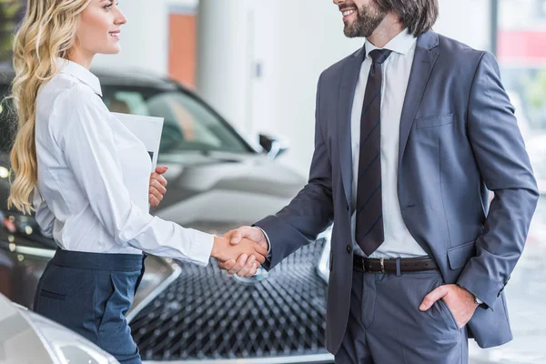 Partial view of auto salon seller and smiling businessman shaking hands at dealership salon — Stock Photo