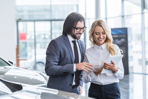 Female auto salon seller with tablet helping smiling businessman to choose car at dealership salon — Stock Photo