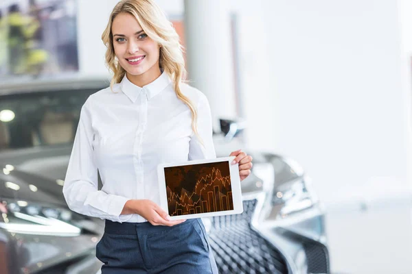 Retrato de vendedor sorrindo mostrando tablet com diagrama gráfico em mãos no salão de concessionárias — Fotografia de Stock