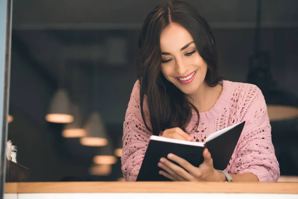 Fröhliche junge Frau macht sich Notizen in Lehrbuch am Tisch im Café — Stockfoto