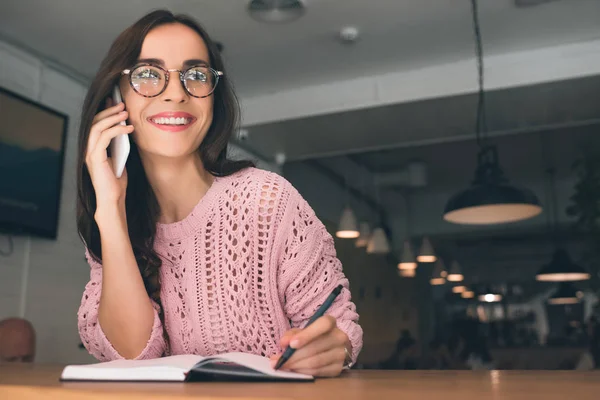 Donna sorridente in occhiali donna che parla su smartphone mentre scrive nel libro di testo a tavola nel caffè — Stock Photo